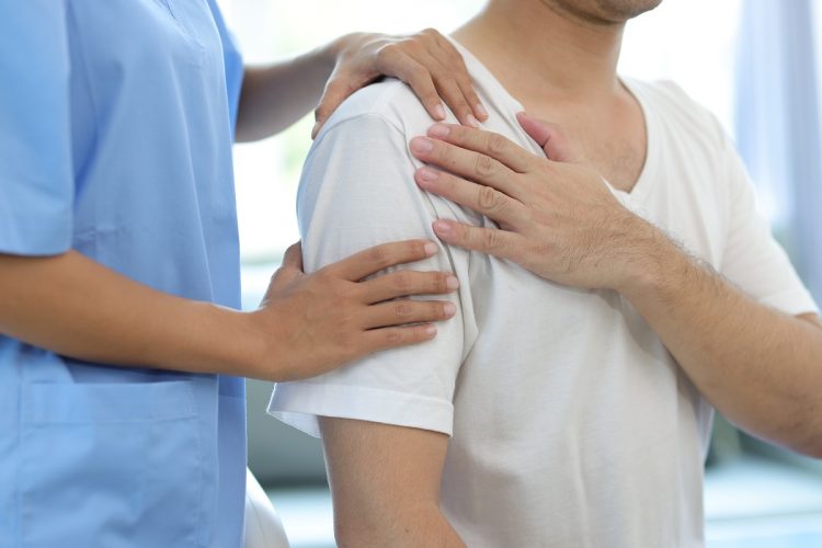 Doctor diagnosing man’s shoulder pain in hospital examination room. Nurse doing massage and physiotherapy to treat patient’s shoulder.